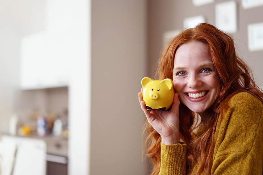 Redheaded woman shows off her yellow piggy bank while relaxing in her kitchen, enjoying her new smile. When considering cosmetic dentistry think about more than the just the cost!