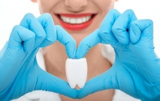 Attractive female dentist placing her hands in a heart shape while holding an oversized tooth. Brushing your teeth and having a routine visit to your dentist can reduce your heart risks.