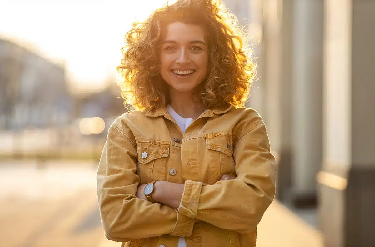 Young woman with wavy golden hair wearing a tan jean jacket, folding her arms, showcasing her improved smile